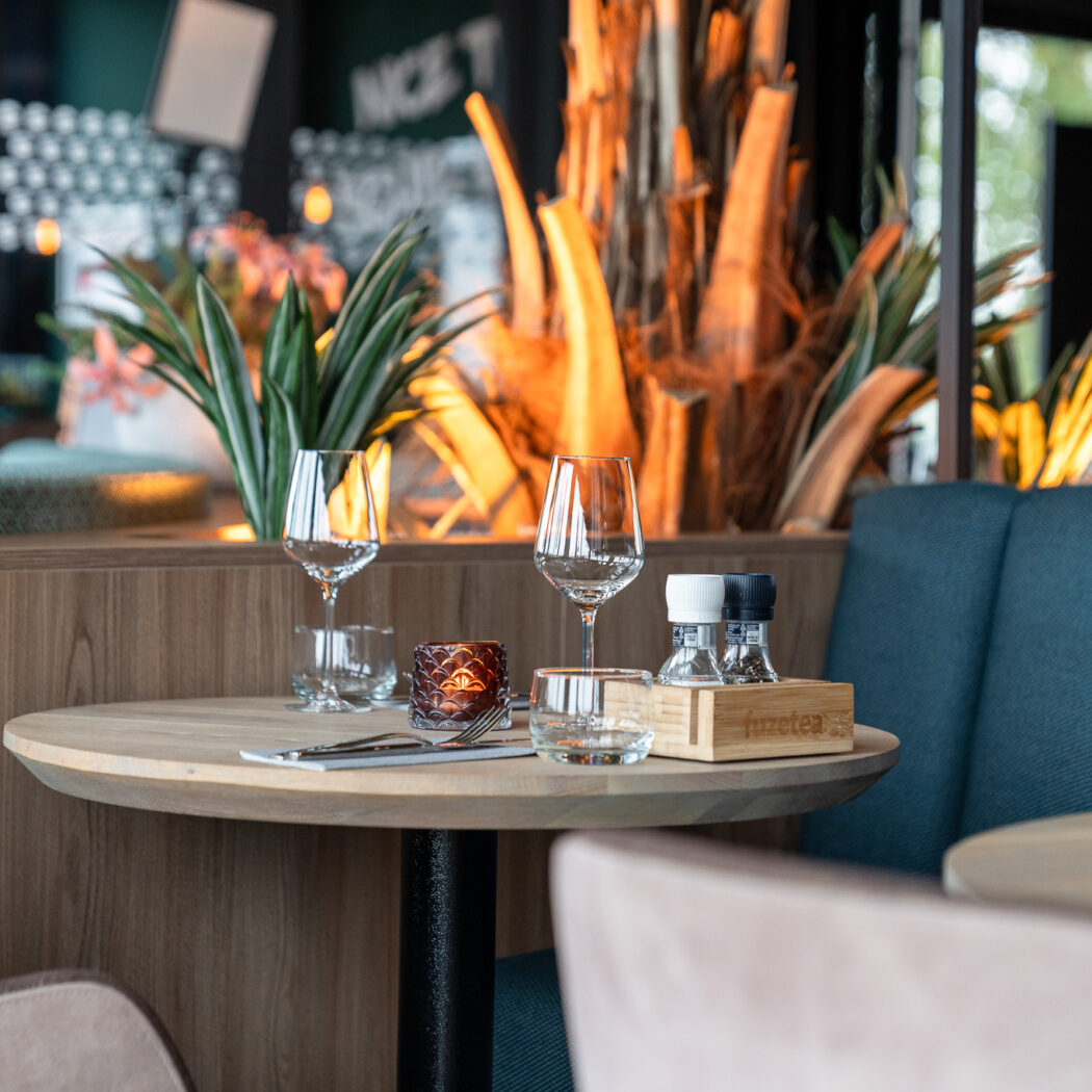 Gedekte houten tafel in restaurant met glazen, bestek, kaars en fuzetea zout- en peperstel.