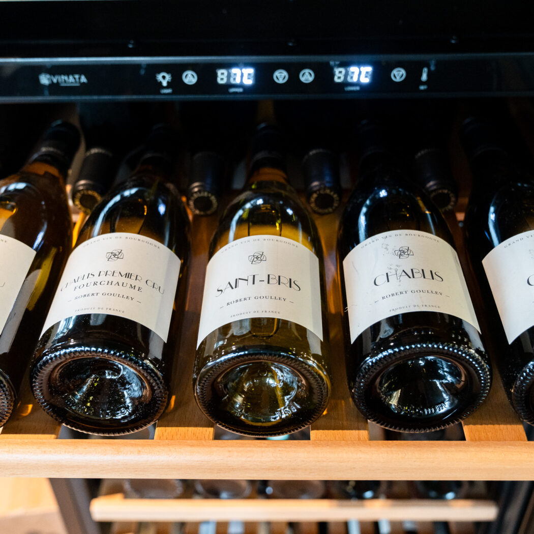Horizontaal liggende wijnflessen met labels Chablis en Saint-Bris op houten rekken in een Vinata wijnkoelkast, 8.3°C.