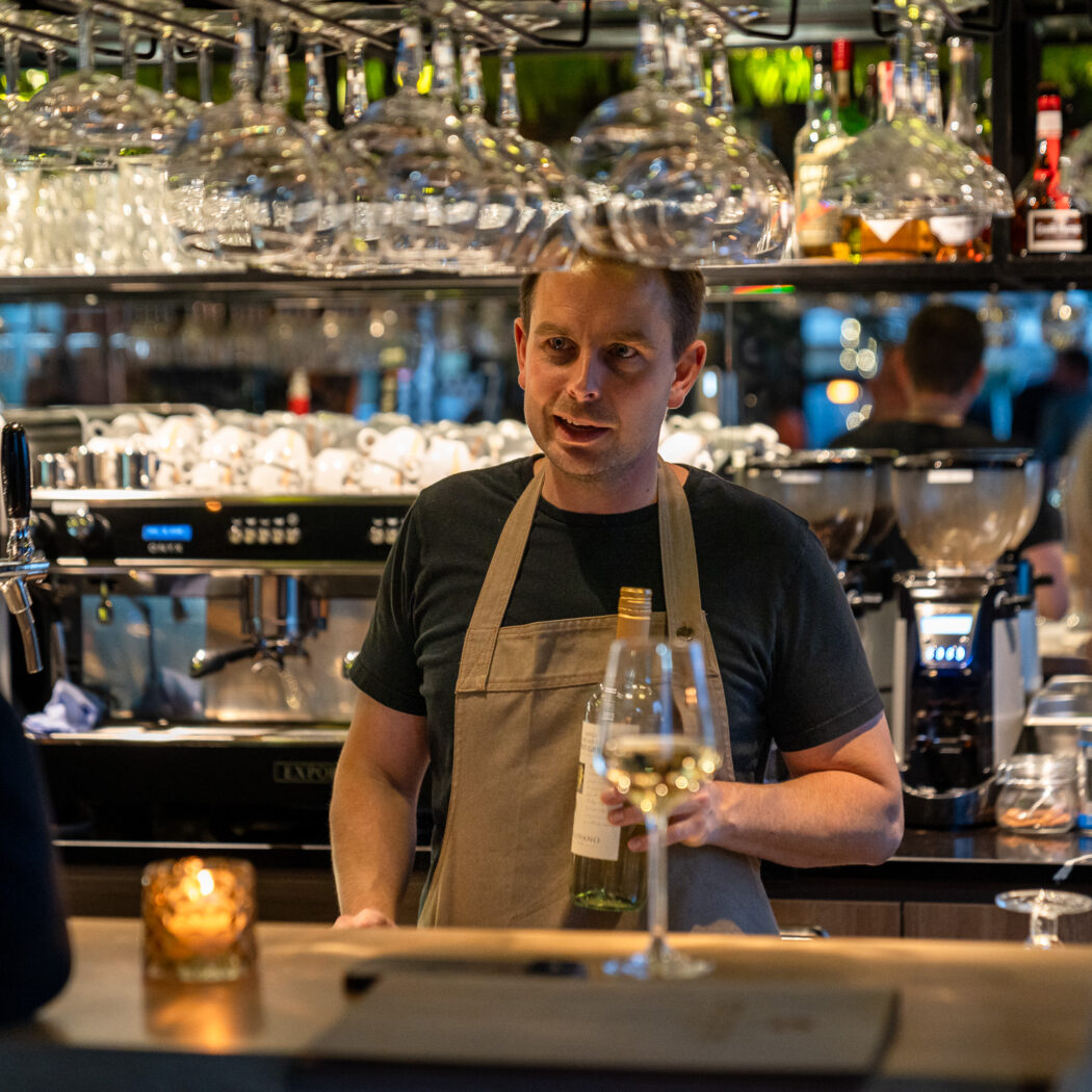 Barman met schort in gesprek met klant, met een fles witte wijn en koffiezetapparaten in de achtergrond.