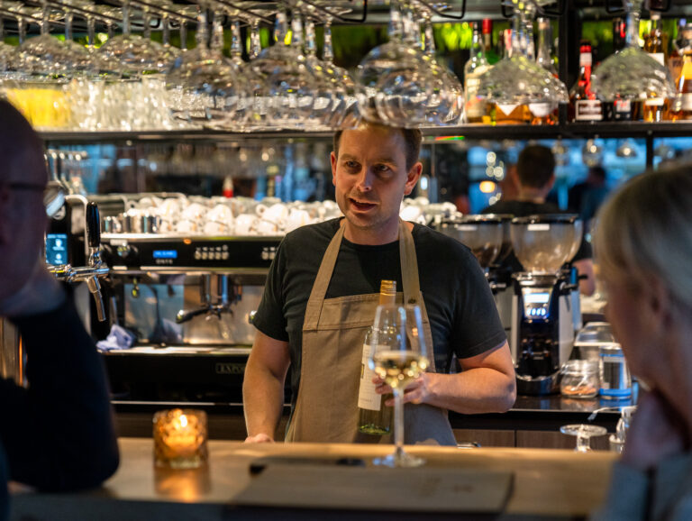 Barman met schort in gesprek met klant, met een fles witte wijn en koffiezetapparaten in de achtergrond.