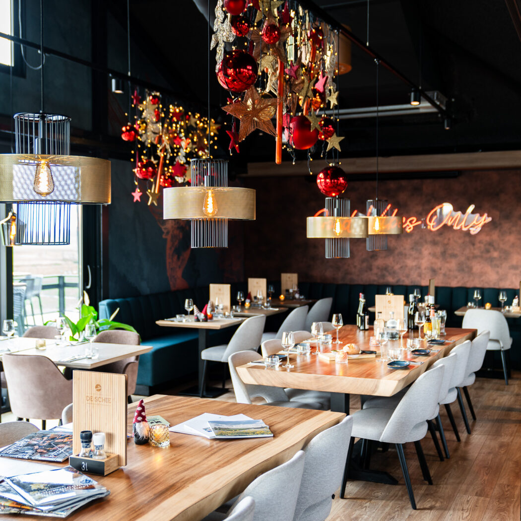 Sfeervol restaurant met kerstversiering, moderne lampen, houten tafels, Its Only neon en terrasuitzicht.