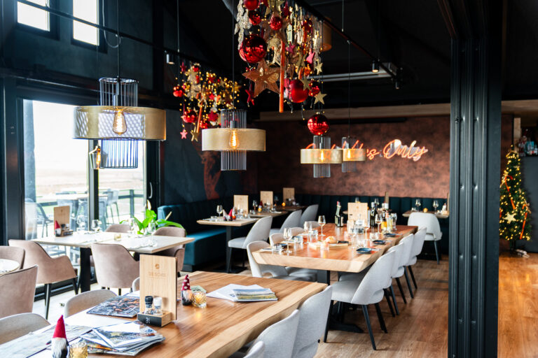 Sfeervol restaurant met kerstversiering, moderne lampen, houten tafels, Its Only neon en terrasuitzicht.