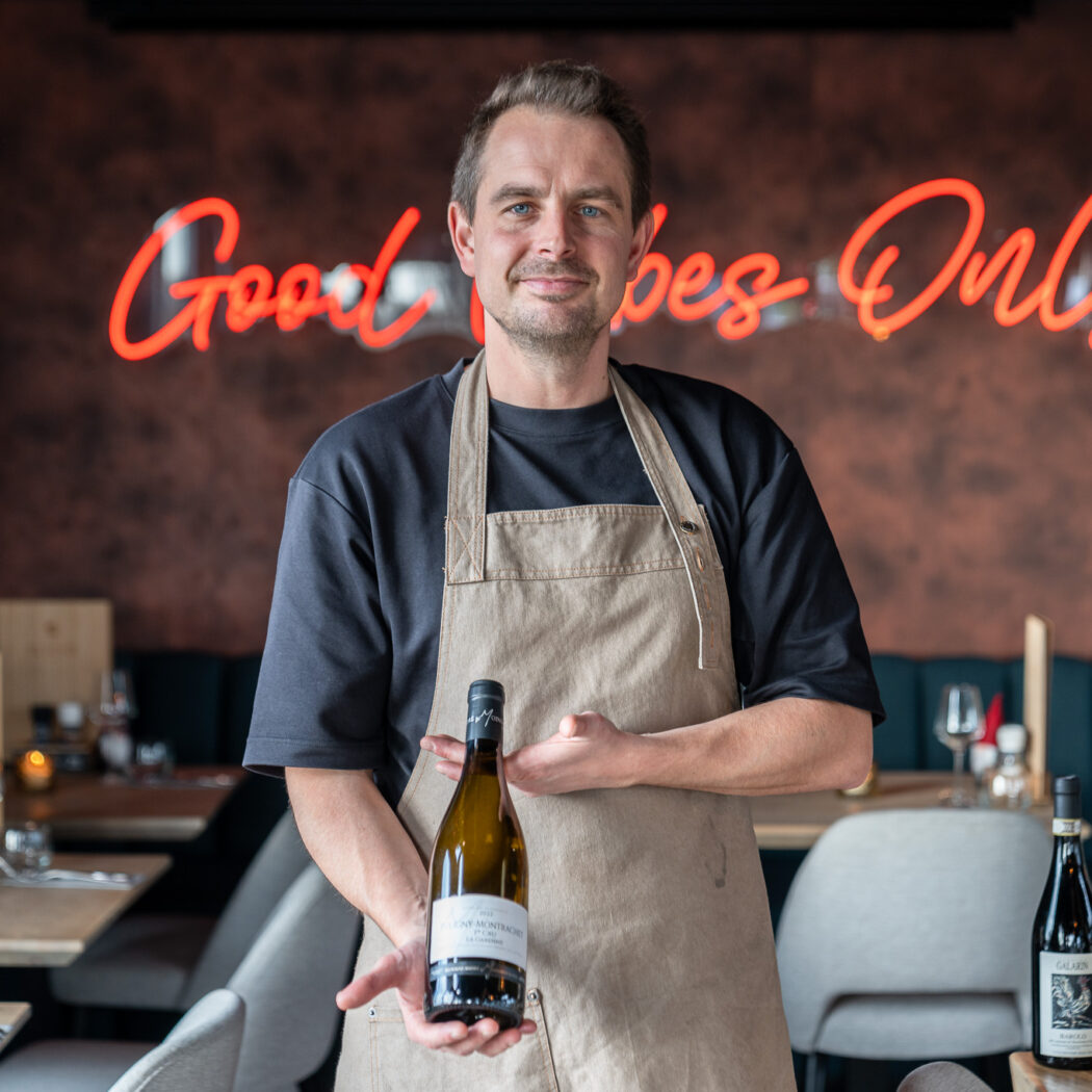 Man in beige schort toont fles witte wijn SAINT-MONTRACHET in restaurant met Good Vibes Only neonlicht.