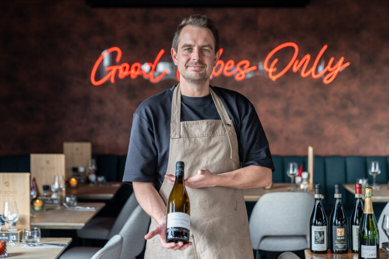 Man in beige schort toont fles witte wijn SAINT-MONTRACHET in restaurant met Good Vibes Only neonlicht.