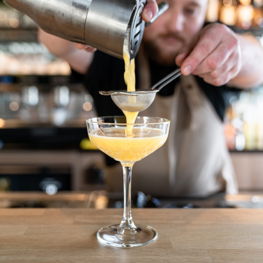 Een barman zeeft een gele cocktail uit een shaker in een coupe glas aan de houten bar.