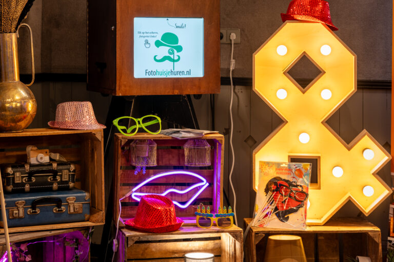 Fotobooth met scherm Fotohuisjehuren.nl, omringd door brillen, hoeden, een neon lipsign en een verlicht ampersand.