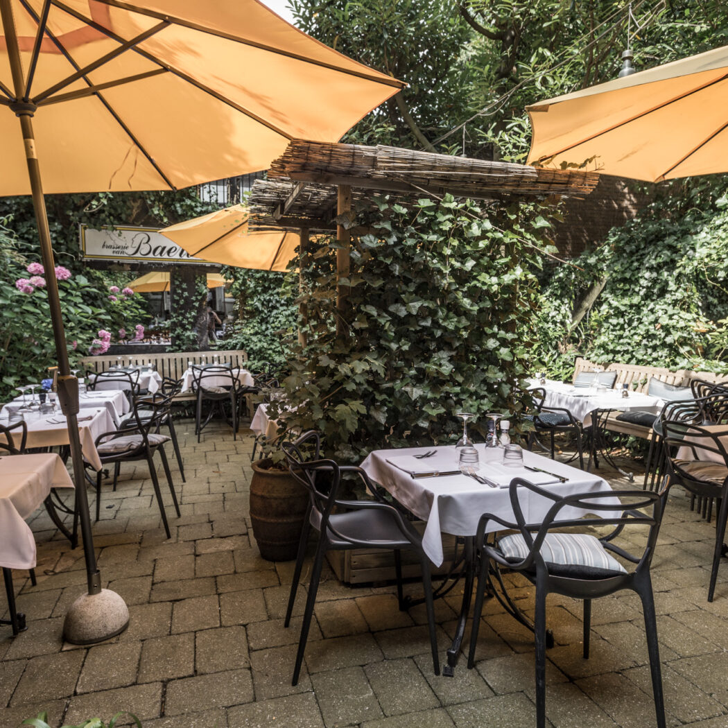 Buiten terras van brasserie van Baer met witgedekte tafels, zwarte stoelen, gele parasols en weelderige groene planten.