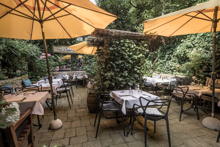 Buiten terras van brasserie van Baer met witgedekte tafels, zwarte stoelen, gele parasols en weelderige groene planten.