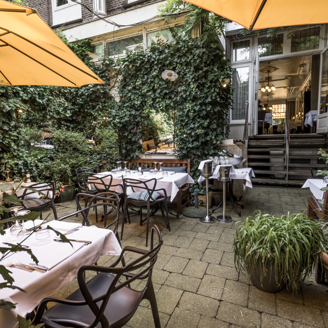 Groen restaurant terras met gedekte tafels onder gele parasols, leidend naar de ingang van het gebouw.