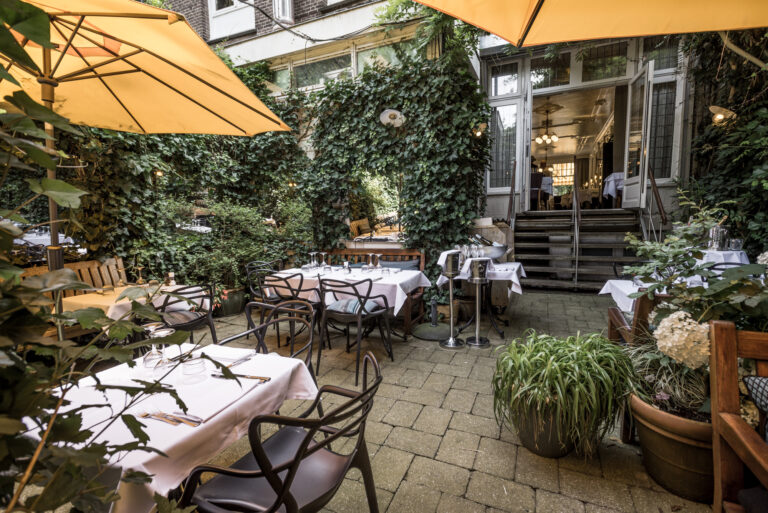 Groen restaurant terras met gedekte tafels onder gele parasols, leidend naar de ingang van het gebouw.