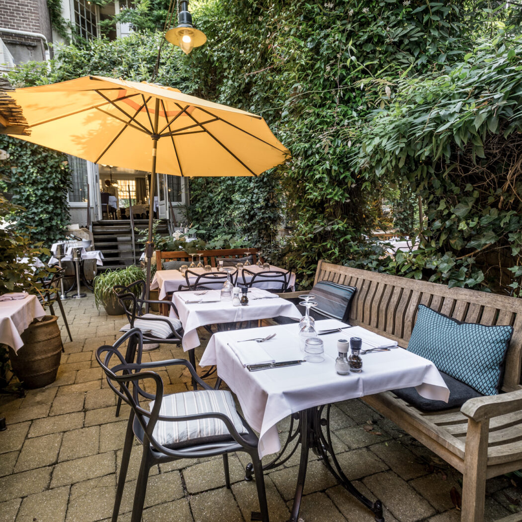 Sfeervol buitenterras van een restaurant met gedekte tafels, houten bank en een grote gele parasol omgeven door groen.