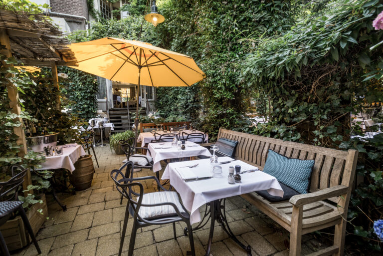 Sfeervol buitenterras van een restaurant met gedekte tafels, houten bank en een grote gele parasol omgeven door groen.