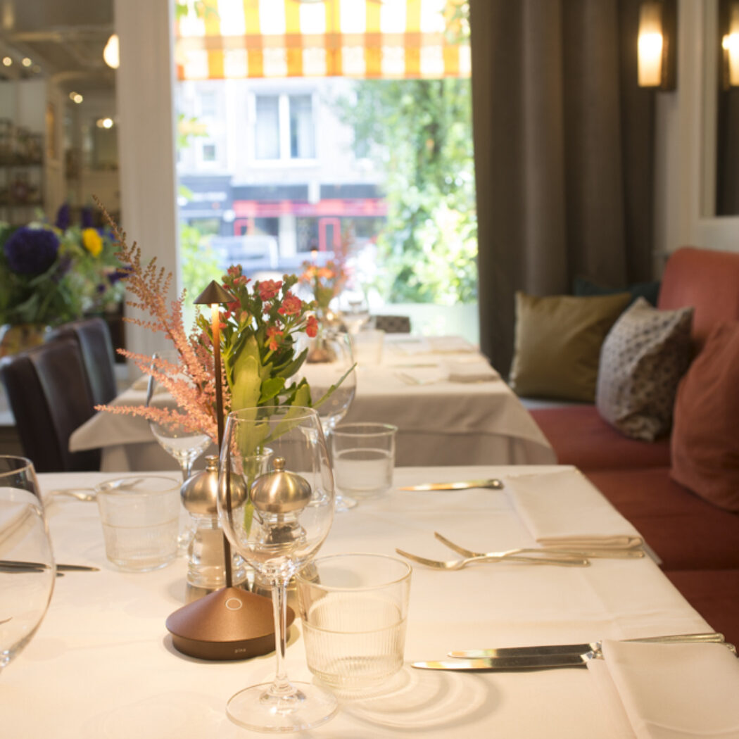 Elegant restaurantinterieur met gedekte tafels, bloemstukken en oranje banken bij een raam met gestreept zonnescherm.