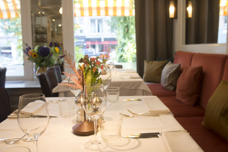 Elegant restaurantinterieur met gedekte tafels, bloemstukken en oranje banken bij een raam met gestreept zonnescherm.