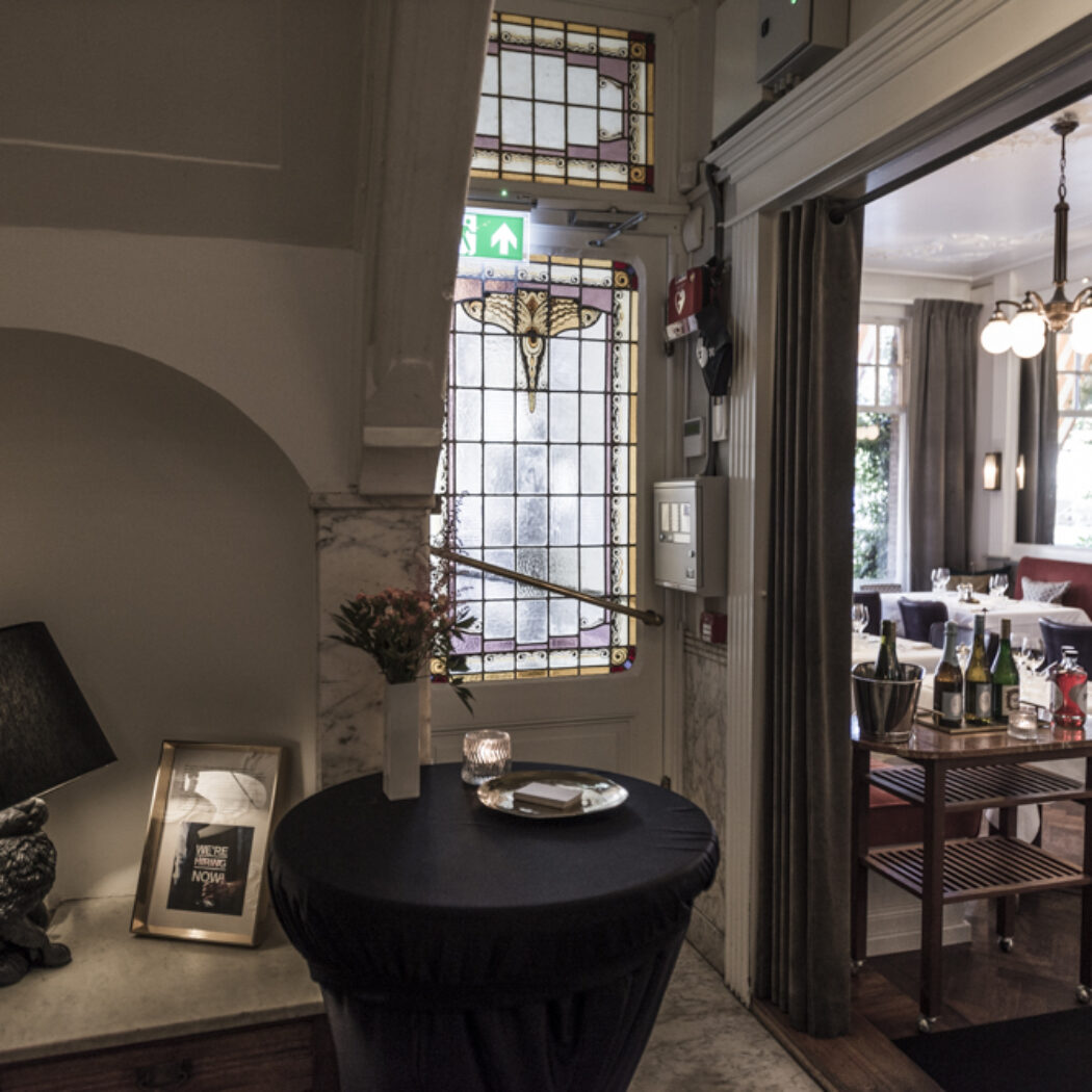 Gang met konijnenlamp en glas-in-looddeur, die uitkomt op een restaurant met gedekte tafels en drankkar.