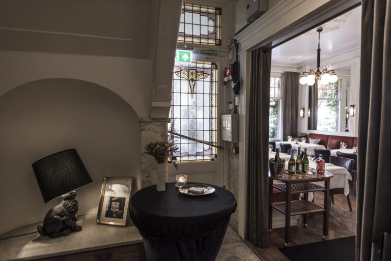 Gang met konijnenlamp en glas-in-looddeur, die uitkomt op een restaurant met gedekte tafels en drankkar.