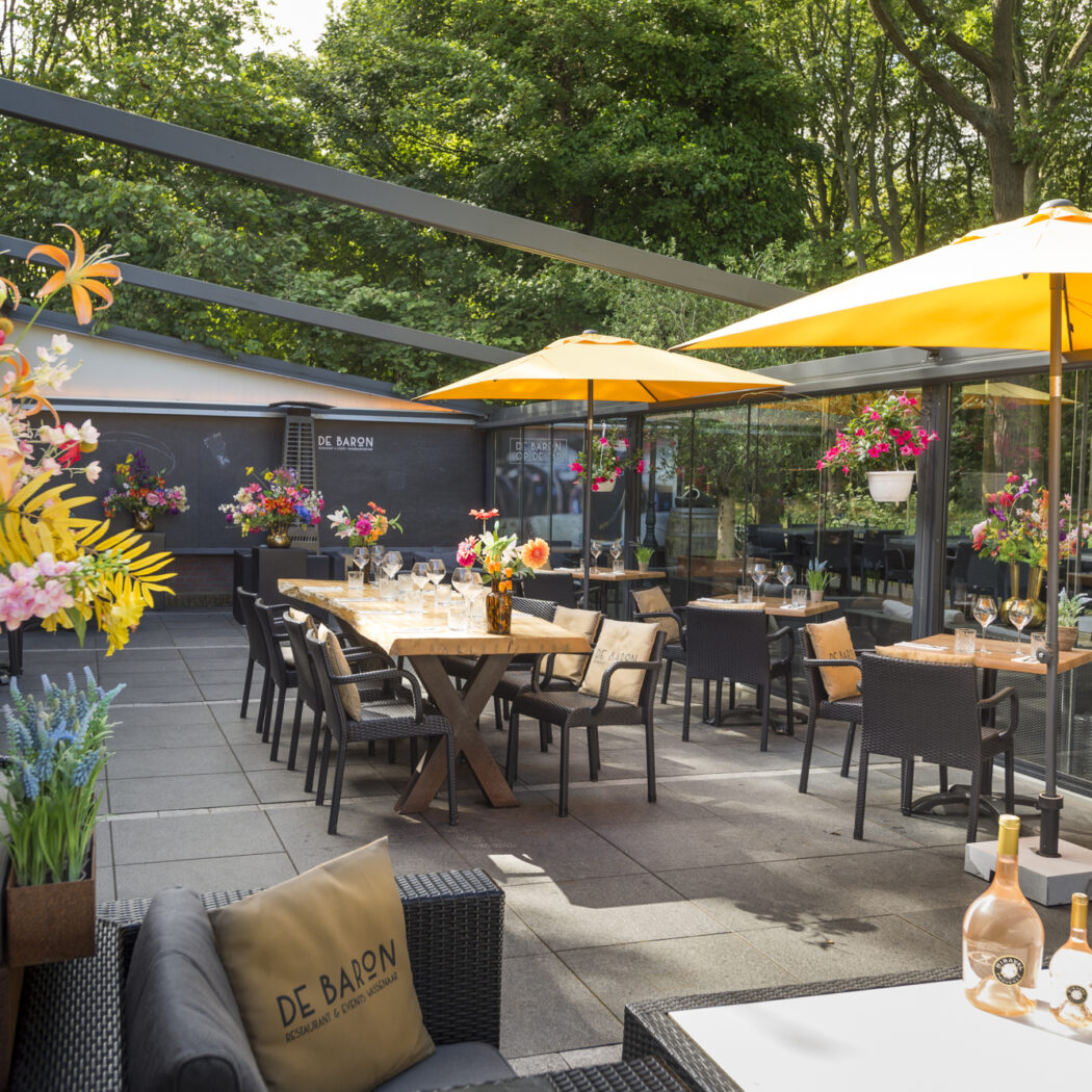 Zonnig buitenterras van restaurant De Baron met gedekte tafels, zwarte stoelen, gele parasols en weelderige bloemstukken.