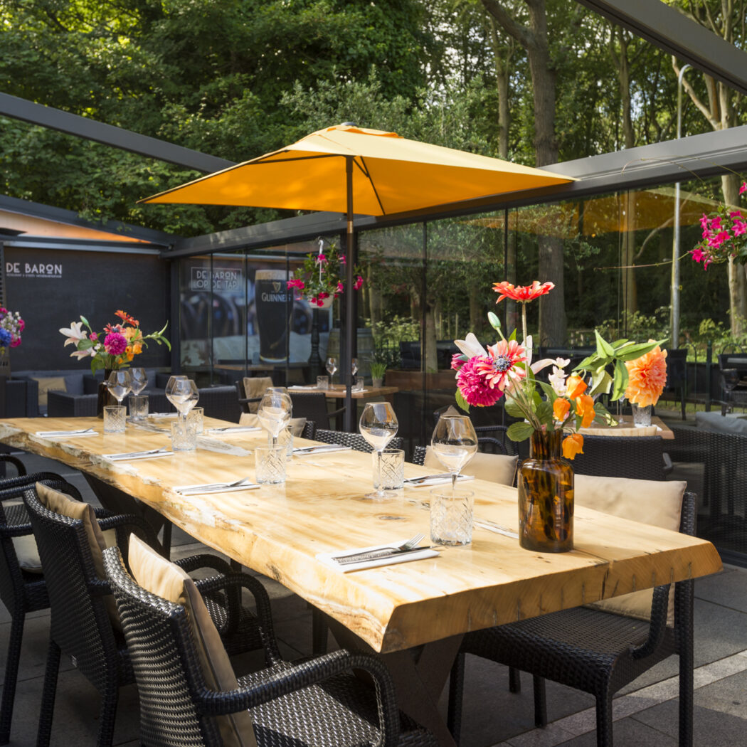 Gezellig restaurantterras van De Baron met lange houten tafel, zwarte stoelen, bloemen en oranje parasol.