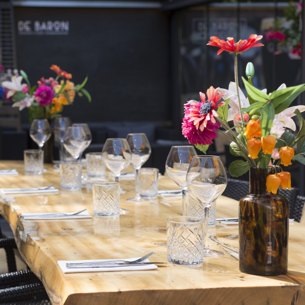 Een gedekte, lange houten terrastafel met diverse glazen, bestek en kleurrijke bloemdecoraties op het terras van DE BARON.