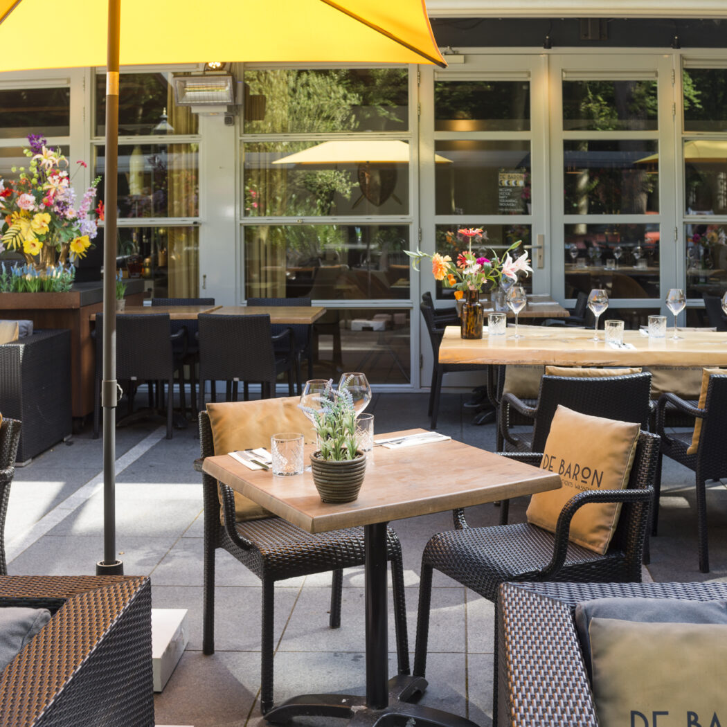 Zonnig terras van restaurant De Baron met houten tafels, rotan stoelen en gele parasols.