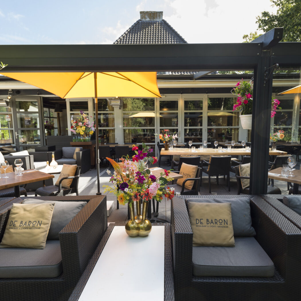 Gezellig buitenterras van restaurant De Baron met loungebanken, eettafels, gele parasols en kleurrijke bloemen.