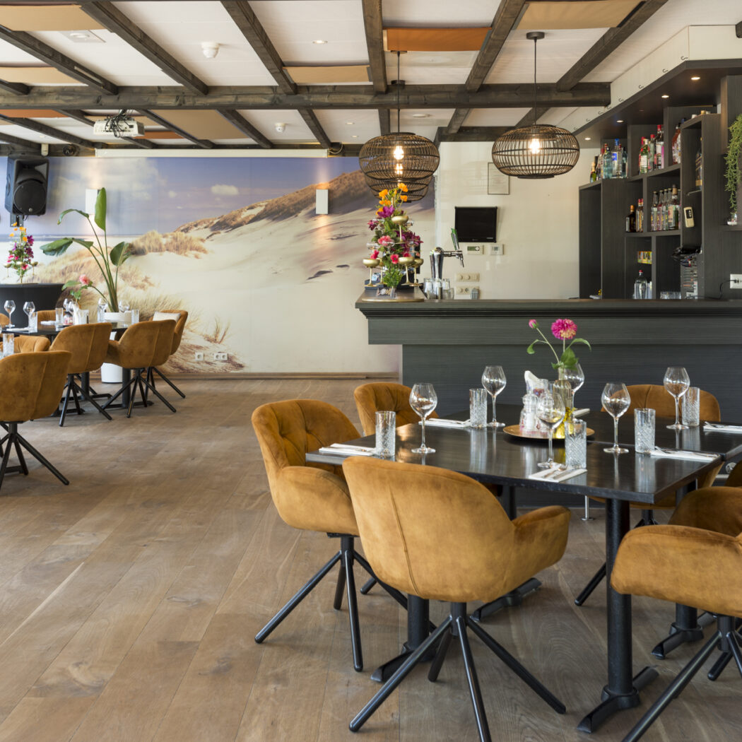 Ruim restaurant met comfortabele cognac stoelen, donkere tafels, een bar en een wandafbeelding van strandduinen.