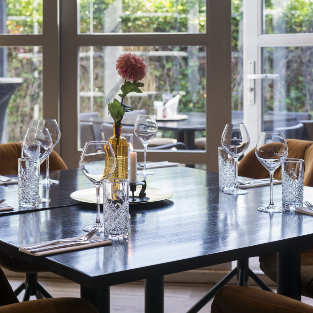 Gedekte donkere eettafel in restaurant met bruine stoelen, wijnglazen met Bachstad logo, waterglazen, kaars en roze bloem.