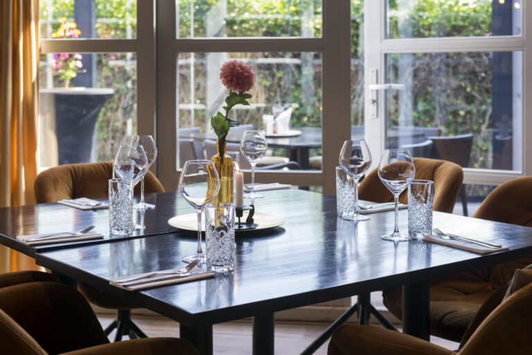 Gedekte donkere eettafel in restaurant met bruine stoelen, wijnglazen met Bachstad logo, waterglazen, kaars en roze bloem.