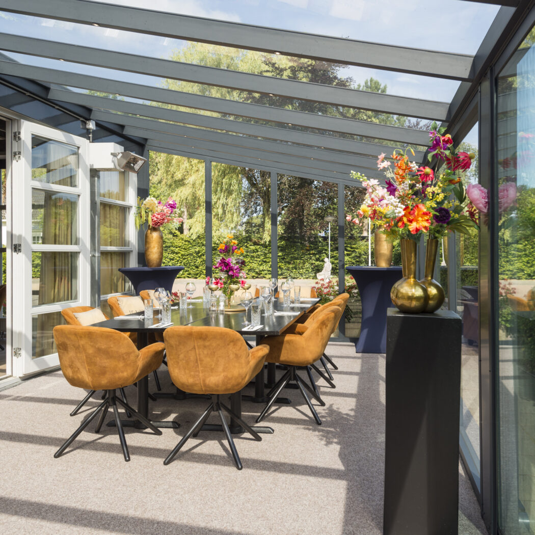 Lichte glazen serre met gedekte eettafel, comfortabele oranje stoelen en kleurrijke bloemstukken in gouden vazen.
