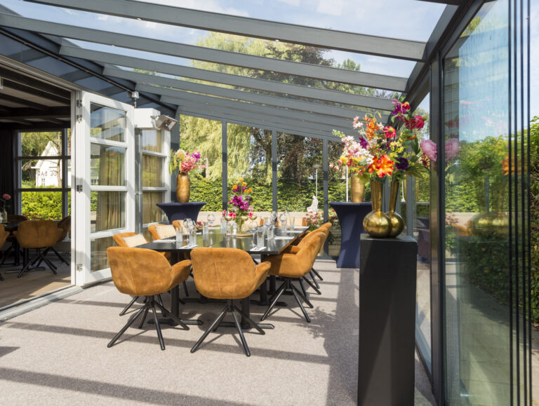 Lichte glazen serre met gedekte eettafel, comfortabele oranje stoelen en kleurrijke bloemstukken in gouden vazen.