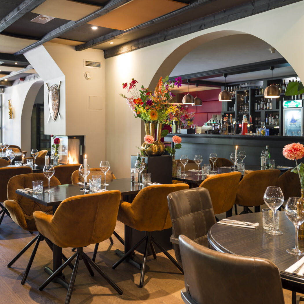 Sfeervol restaurantinterieur met gedekte tafels, comfortabele bruine en oranje stoelen, een bar en een open haard.