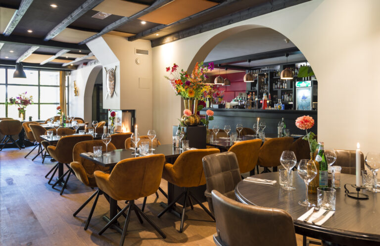 Sfeervol restaurantinterieur met gedekte tafels, comfortabele bruine en oranje stoelen, een bar en een open haard.