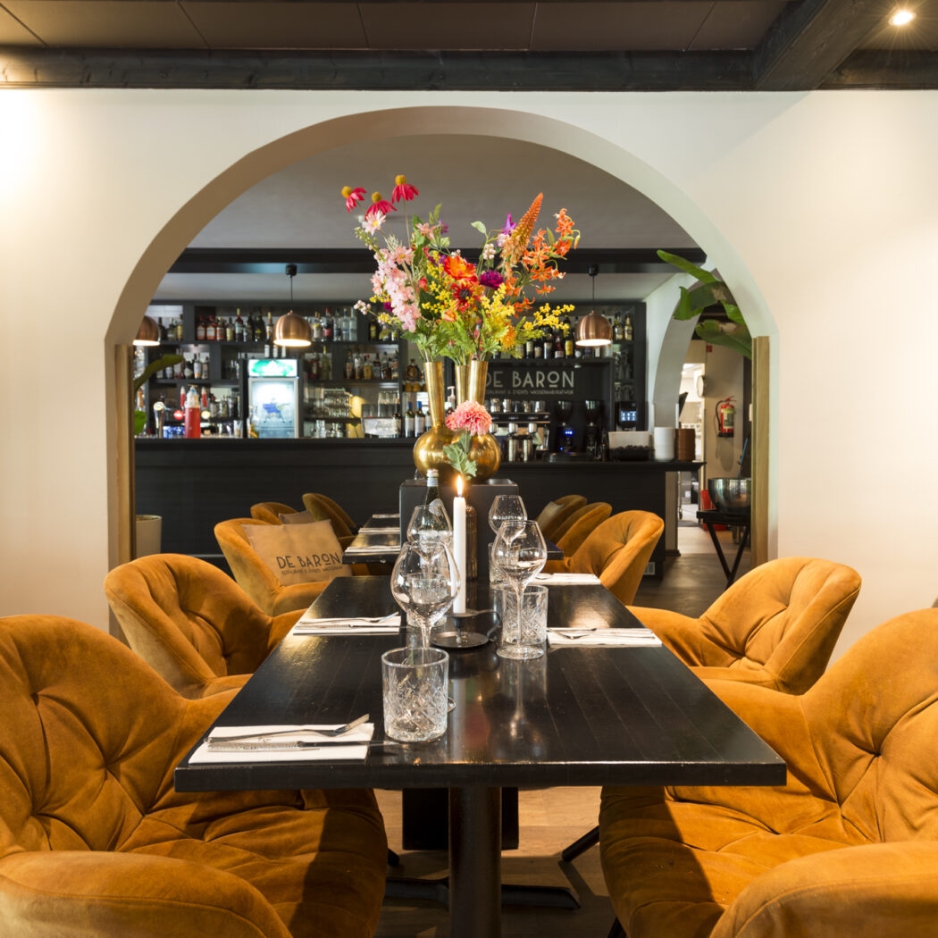 Sfeervolle restaurant eetzaal met oranje fluwelen stoelen, donkere tafels met bloemstuk en bar DE BARON door een boog.