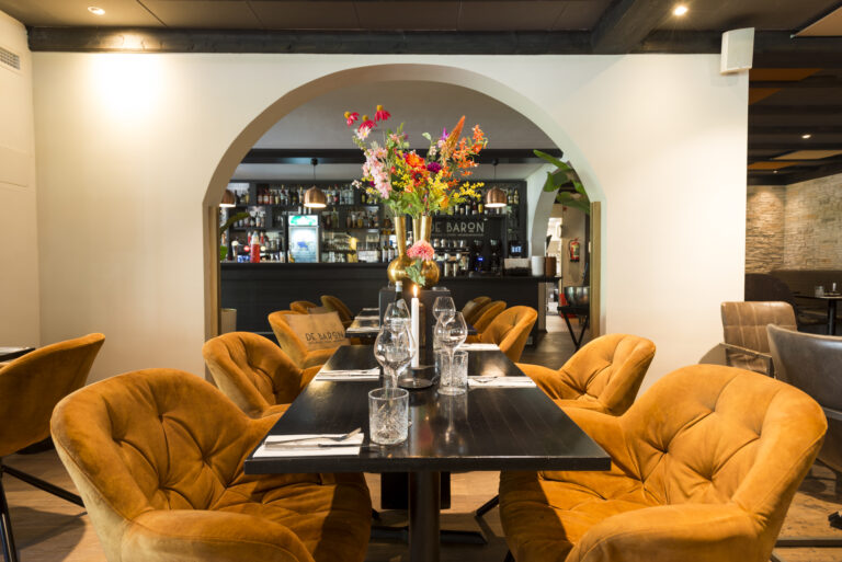Sfeervolle restaurant eetzaal met oranje fluwelen stoelen, donkere tafels met bloemstuk en bar DE BARON door een boog.