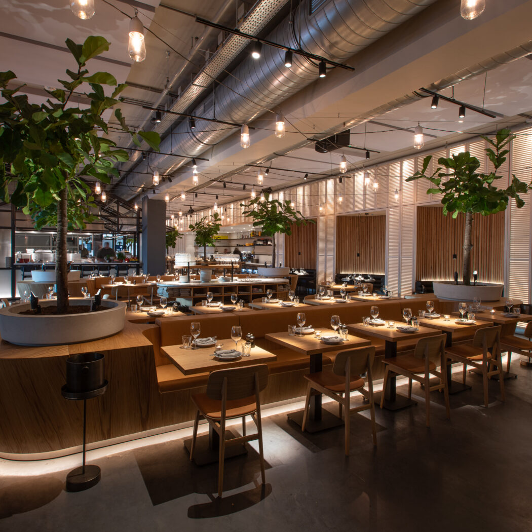 Modern restaurant met houten tafels en banken, grote potplanten en industriële verlichting, gedekt voor gasten.
