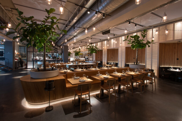 Modern restaurant met houten tafels en banken, grote potplanten en industriële verlichting, gedekt voor gasten.