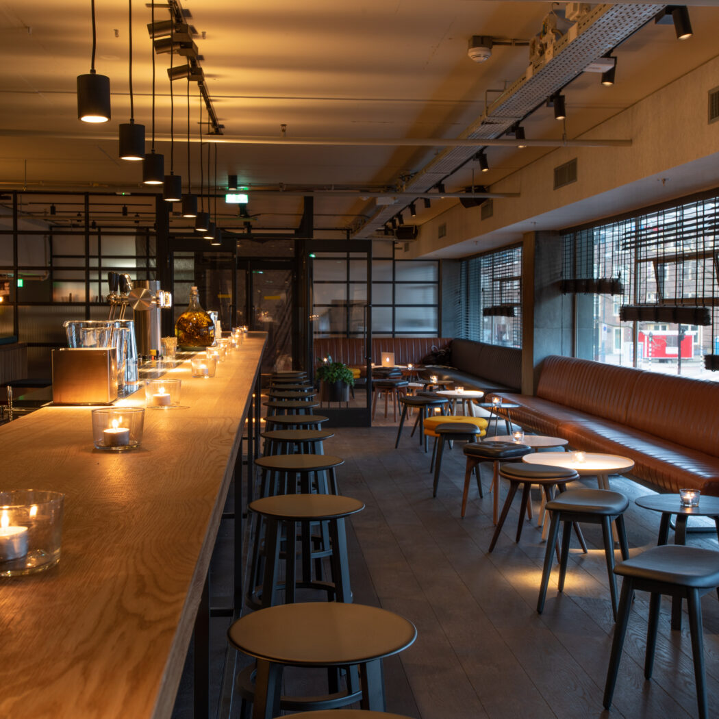 Sfeervol barinterieur met lange houten bar, zwarte krukken en een bruine lederen bank, verlicht door kaarslicht.