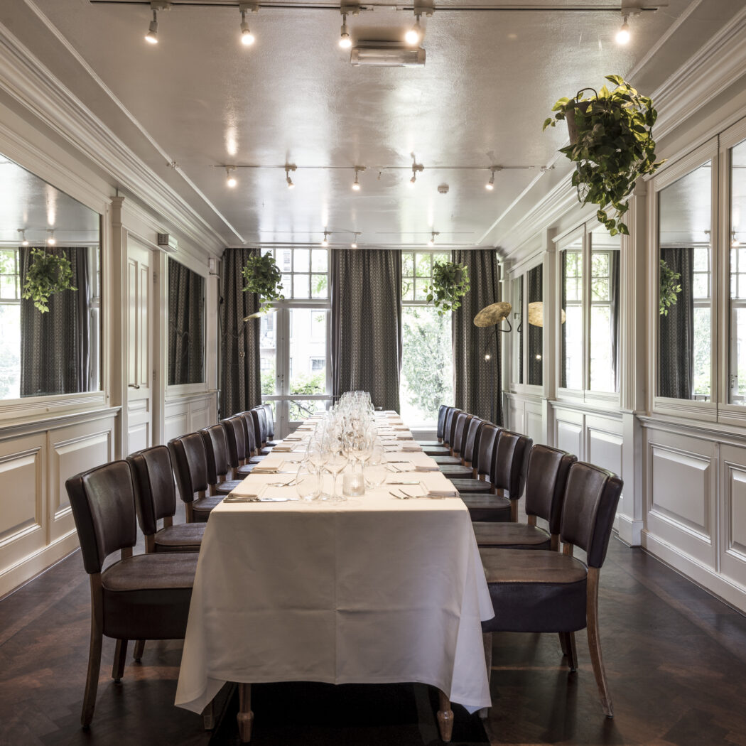 Lange gedekte tafel met lederen stoelen in een klassieke dinerzaal met spiegels en hangplanten.