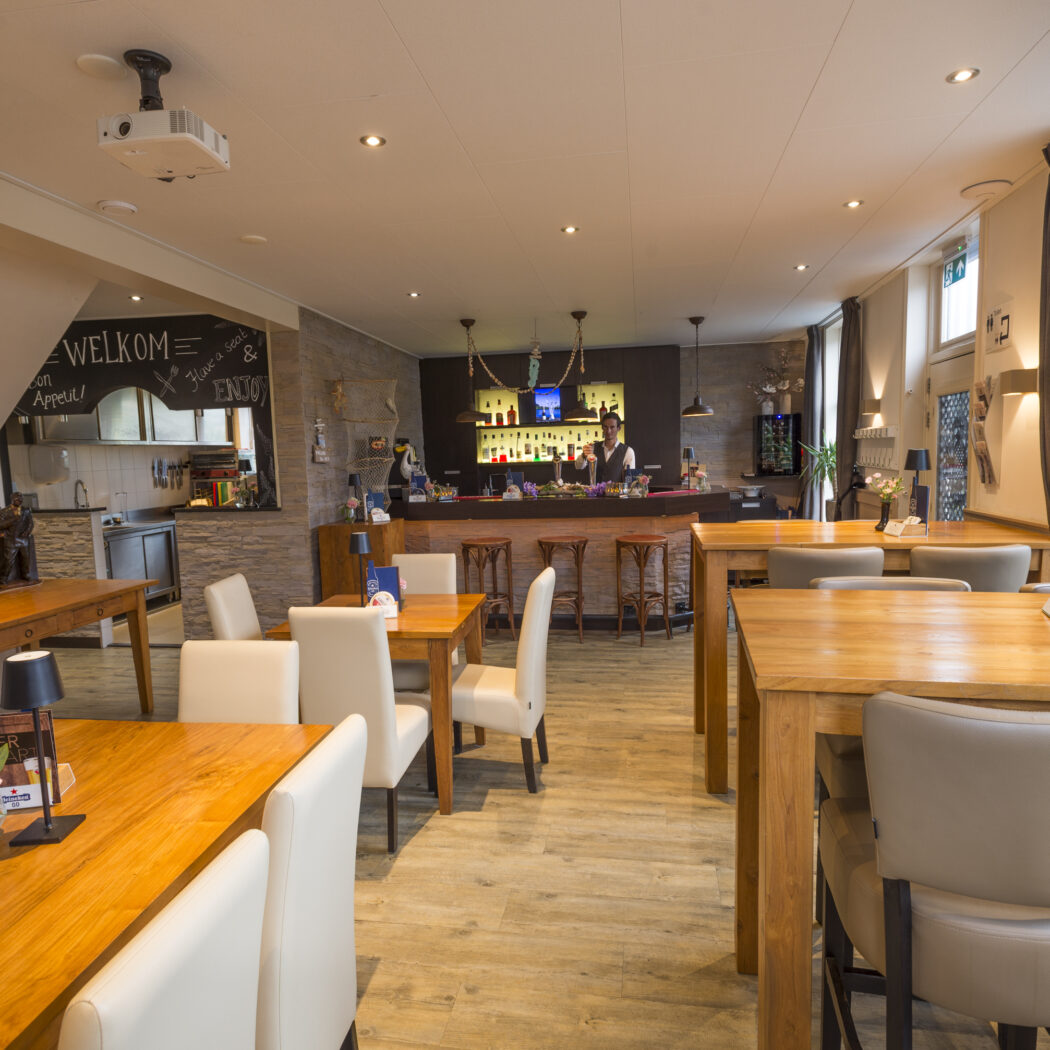 Sfeervol restaurantinterieur met houten tafels, diverse stoelen, een bar met barman en een krijtmuur met WELKOM.