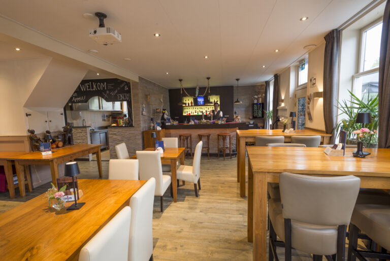 Sfeervol restaurantinterieur met houten tafels, diverse stoelen, een bar met barman en een krijtmuur met WELKOM.