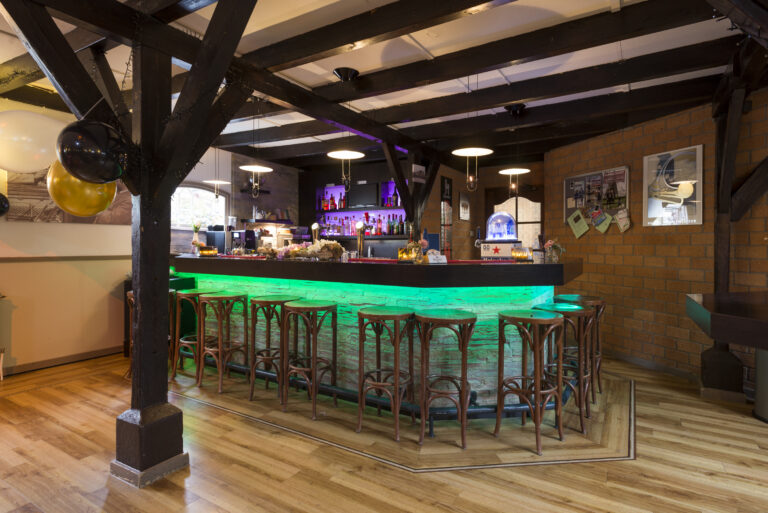 Bar in evenementenlocatie met groene LED-verlichting, houten krukken, donkere balken en feestelijke ballonnen.