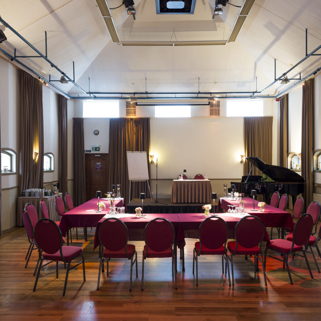 Klassieke conferentiezaal met houten vloer, rode tafels, stoelen, podium met scherm en zwarte vleugel.