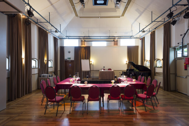 Klassieke conferentiezaal met houten vloer, rode tafels, stoelen, podium met scherm en zwarte vleugel.