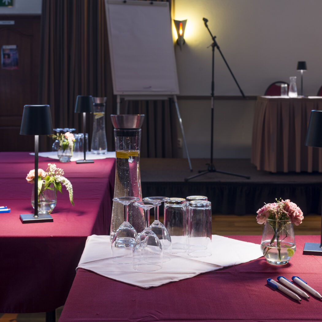 Gedekte bordeauxrode tafels met water, glazen, pennen, lampen en bloemen in een vergaderzaal, podium achterin.