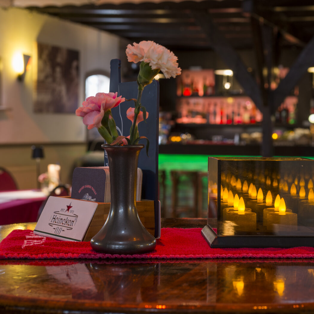 Ronde restauranttafel met rode tafelloper, vaas roze anjers, Heineken onderzetters en een lantaarn met LED kaarsen.