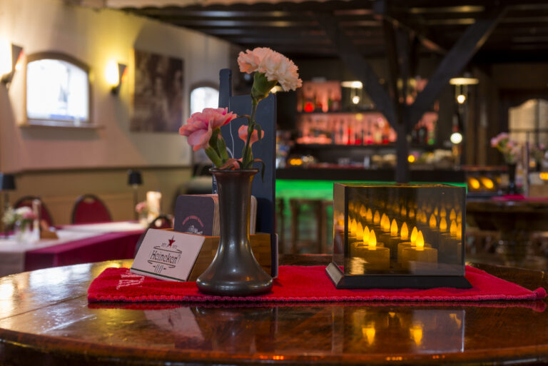 Ronde restauranttafel met rode tafelloper, vaas roze anjers, Heineken onderzetters en een lantaarn met LED kaarsen.