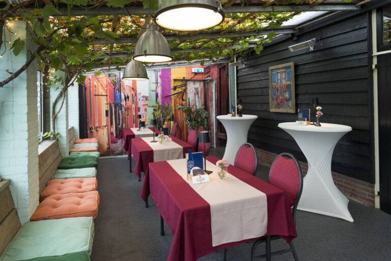 Gezellig overdekt terras met druivenranken, eettafels met rode kleden en een muurprint van een kleurrijke straat.