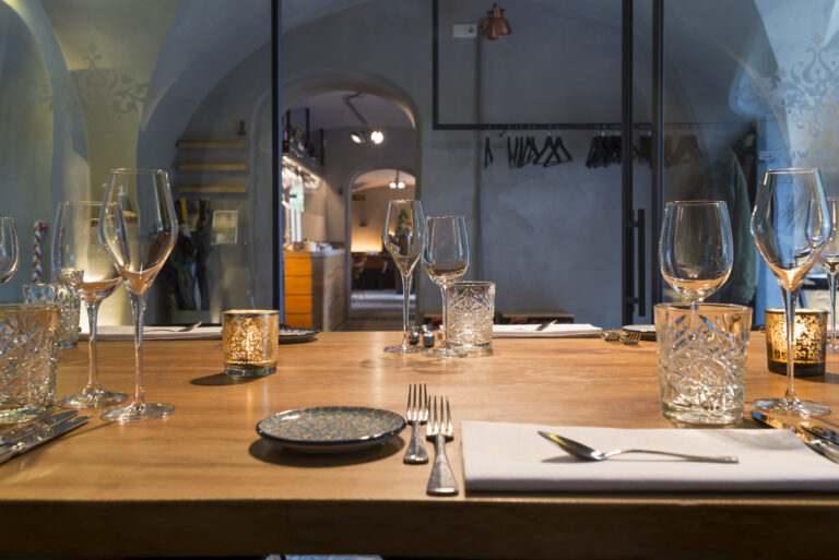 Sfeervolle gedekte houten tafel in een restaurant, met wijnglazen, bestek, en brandende kaarslichtjes.