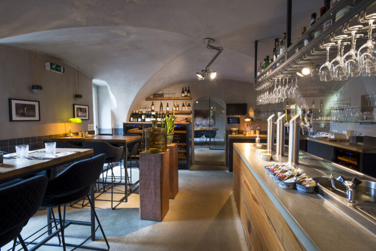 Sfeervol restaurantinterieur met houten bar, bierkranen, wijnglazen en zitjes onder boogplafond.