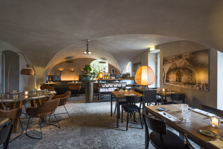 Sfeervol restaurantinterieur met gewelfde plafonds, houten tafels, koperen vloerlamp en rieten tafellamp.